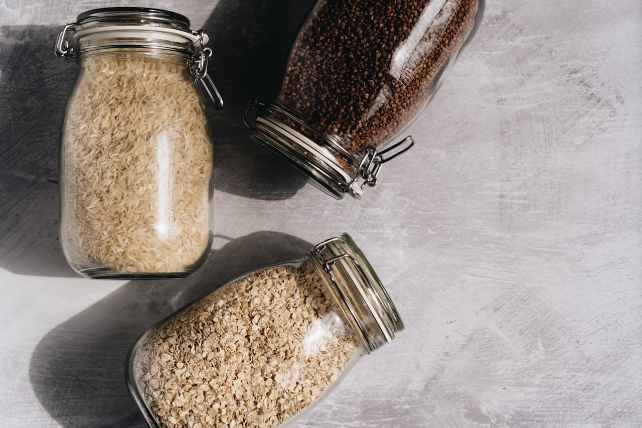 Top-down view of rice, oats, and buckwheat in glass jars on a textured surface.
