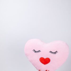 A hand holds a cute heart-shaped pillow featuring a lovely face and red heart lips, perfect for Valentine's Day.