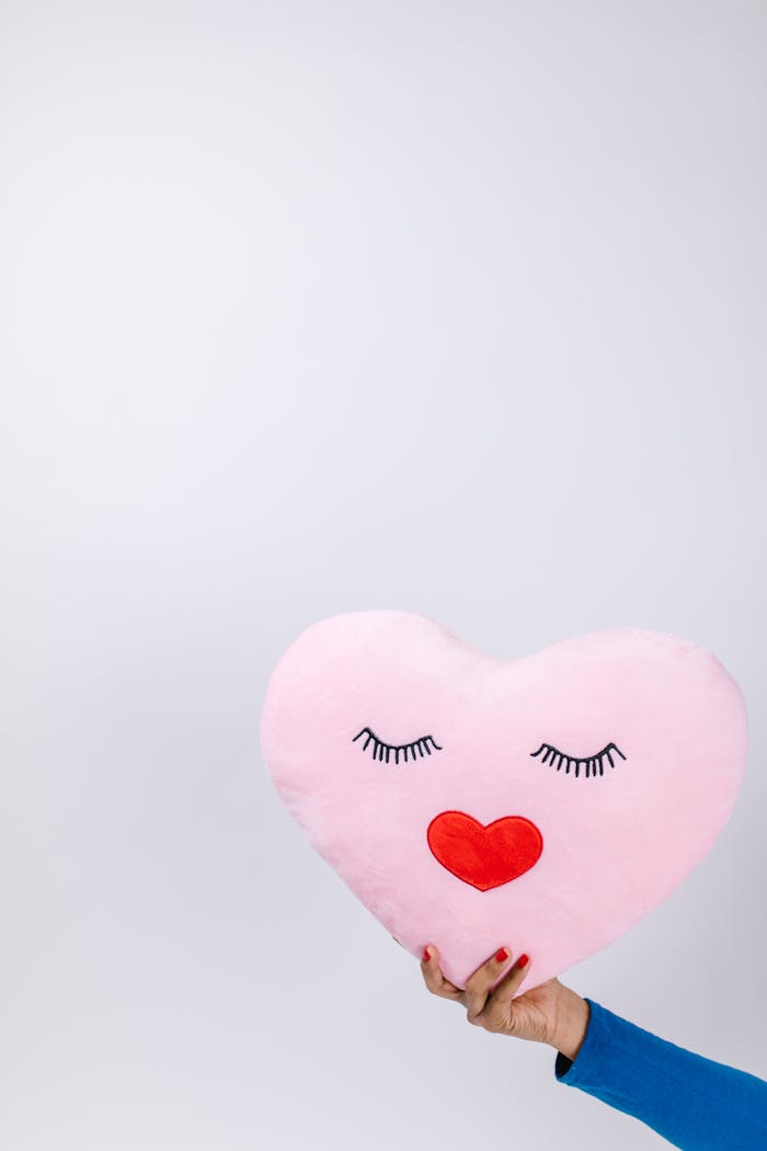 A hand holds a cute heart-shaped pillow featuring a lovely face and red heart lips, perfect for Valentine's Day.