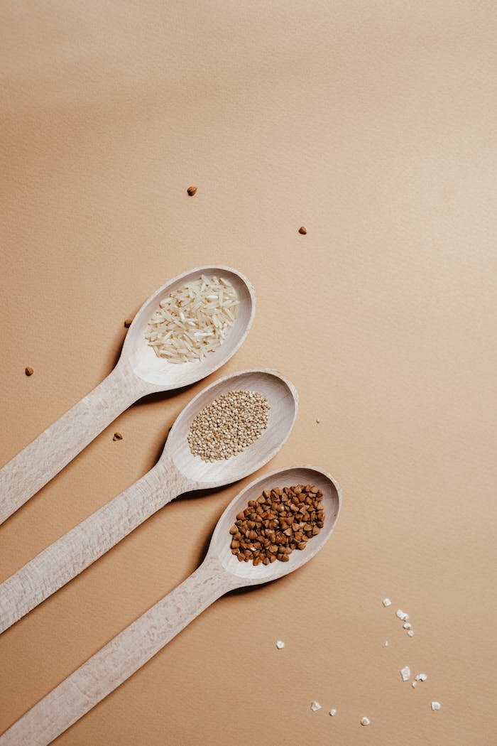 Top view of rice, quinoa, and buckwheat in wooden spoons on neutral background.